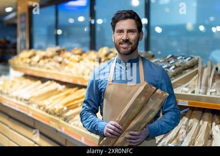 Bäckerei-Angestellter lächelt, während er frische Baguettes hält. Brotschnitt im Hintergrund mit verschiedenen Brottypen. Konzept von freundlichem Kundenservice und frischen Backwaren Stockfoto