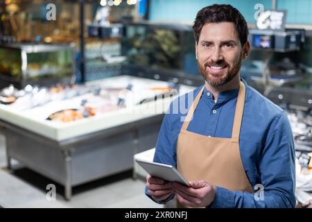 Lächelnder Fischhändler mit Schürze und Tablette auf dem modernen Fischmarkt. Frischer Fisch und Meeresfrüchte im Hintergrund. Bild vermittelt Professionalität, Technologie, frische Lebensmittel, Geschäfte und Service. Stockfoto