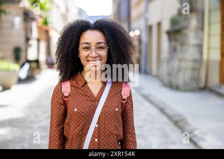 Eine junge Frau mit lockigem Haar und Brille, lässig mit einem Rucksack gekleidet, lächelt, während sie durch eine malerische Straße schlendert und die Freude an Reisen und Abenteuer fängt. Stockfoto