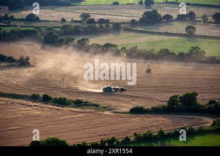 Aus der Vogelperspektive eines Traktors, der auf einem Ackerland arbeitet und Staubwolken erzeugt, umgeben von grünen Feldern und Bäumen in einer ländlichen Landschaft. Stockfoto