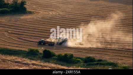 Aus der Vogelperspektive eines Traktors, der während der Erntezeit auf einem riesigen goldenen Feld arbeitet und Staubwolken erzeugt, wenn er sich über das Land bewegt. Stockfoto