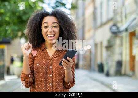 Fröhliche junge Frau, die Erfolg feiert, während sie ihr Smartphone draußen hält. Lächelnde Person, die Glück und Aufregung in urbaner Umgebung ausdrückt. Konzept der Leistung, Positivität und Technologienutzung Stockfoto