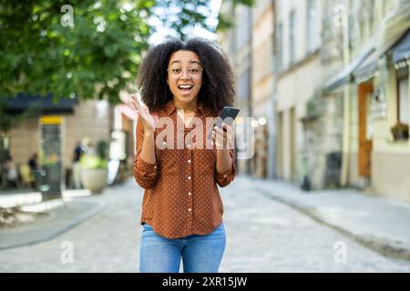 Fröhliche Frau mit lockigem Haar, Brille und lässigem Outfit, hält ein Telefon und feiert einen Sieg im Freien. Sie steht auf einer kopfsteingepflasterten Straße mit Gebäuden und Grün im Hintergrund. Stockfoto
