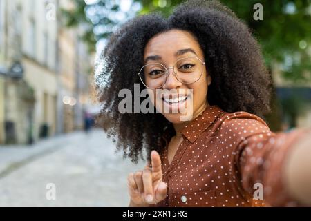 Glückliche Frau lächelt und macht draußen einen Videoanruf mit ihrem Telefon. Sie zeigt auf die Kamera, trägt eine Brille und ein Polka Dot-Shirt, steht auf einer sonnigen Straße. Stockfoto