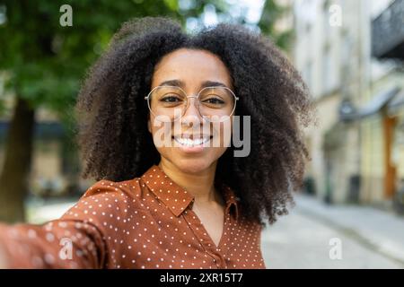 Lebendige Frau mit lockigem Haar und Brille, die an einem sonnigen Tag ein fröhliches Selfie macht. Sie trägt ein Polka Dot-Shirt, das einen offenen und fröhlichen Moment in einer urbanen Outdoor-Umgebung festhält. Stockfoto