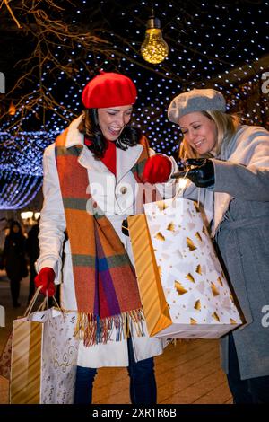 Freundinnen in Mänteln und französische Baskenmützen mit Einkaufstaschen genießen fröhliche Weihnachtseinkäufe auf einem festlich geschmückten Stadtplatz. Stockfoto