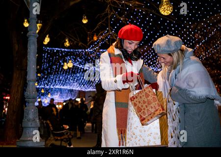 Freundinnen in Mänteln und französische Baskenmützen mit Einkaufstaschen genießen fröhliche Weihnachtseinkäufe auf einem festlich geschmückten Stadtplatz. Stockfoto