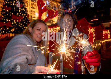 Zwei Freundinnen feiern Silvester auf einem festlich geschmückten Stadtplatz mit beleuchteten Handfunken und Rentiergeweihen. Selektiver Fokus. Stockfoto