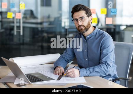 Selbstbewusster Architekt, der an Bauplänen in moderner Büroumgebung arbeitet und sich auf Projektdetails konzentriert. Professionelles Planungsdesign mit Laptop und Notizen. Kreative Arbeitsumgebung Stockfoto