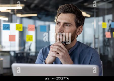 Geschäftsmann im modernen Bürodenken, während er auf Laptop und Stift blickt. Professionelle Reflexion über die Arbeit. Geschäftsumgebung mit farbenfrohen Haftnotizen an der Glaswand. Stockfoto