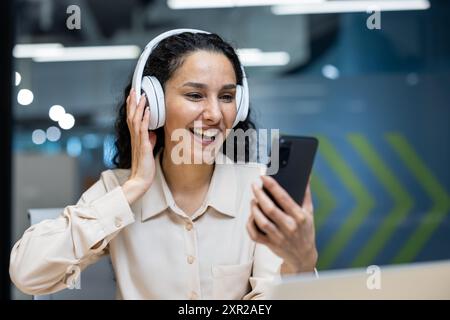 Lächelnde Frau, die während eines Videoanrufs Kopfhörer mit Smartphone trägt, in moderner Büroumgebung. Dame genießt Online-Kommunikation mit dem Telefon, zeigt Glück und positive Emotionen. Stockfoto