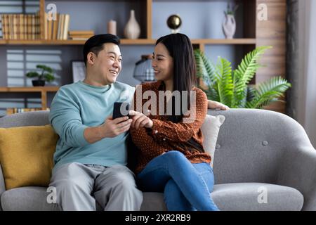 Glückliches asiatisches Paar, das mit dem Smartphone auf der Couch sitzt, die gemeinsame Zeit genießt, über etwas diskutiert oder geteilt wird. Gemütliches Wohnumfeld mit positiven Emotionen und Bindungsmomenten. Stockfoto