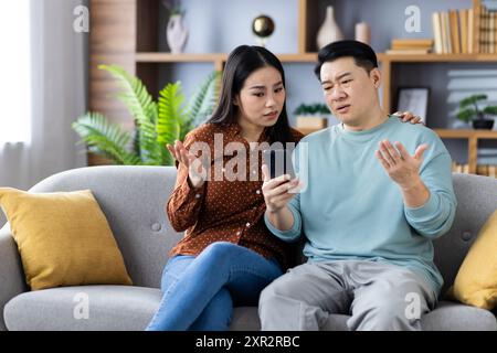 Asiatisches Paar, das auf der Couch sitzt und mit verwirrten Ausdrücken auf das Telefon schaut und über etwas Wichtiges diskutiert. Das Bild zeigt emotionale Interaktion, Kommunikation, Beziehungsdynamik und Technologienutzung Stockfoto
