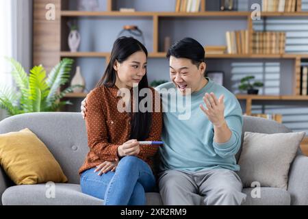 Asiatisches Paar, das auf der Couch sitzt und einen Schwangerschaftstest hält. Mann zeigt Aufregung, während die Frau sich den Test anschaut. Werdende Eltern, die neue Familienmitglieder antizipieren. Stockfoto