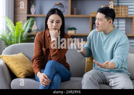 Asiatisches Paar, das auf der Couch eine Diskussion führt. Ehepartner, die Beziehungskonflikte haben. Frau, die aufgebracht aussieht, während der Mann seine Gefühle ausdrückt Stockfoto