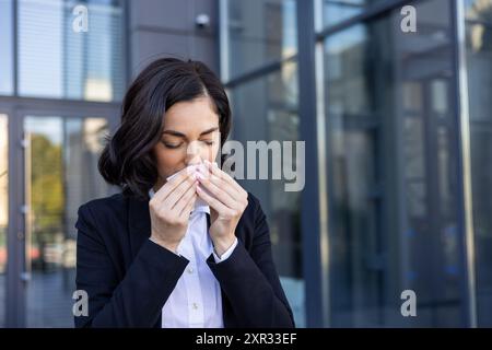 Ein Nahfoto einer kranken jungen Frau in einem Business-Anzug, die vor einem Gebäude steht und sich die Nase abwischt, mit einer Serviette aus einer laufenden Nase. Stockfoto