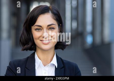 Nahaufnahme eines Porträts einer schönen jungen Frau in einem Business-Anzug, die vor einem Gebäude steht, lächelt und in die Kamera blickt. Stockfoto