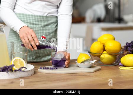 Junge Frau, die Lavendelsaft aus der Flasche in das Glas gießt, um Limonade am Tisch in der Küche zuzubereiten Stockfoto