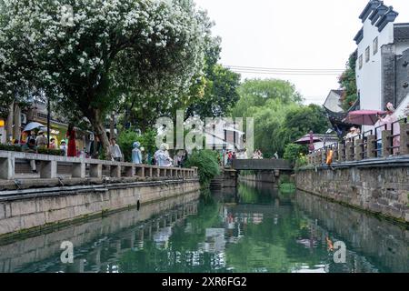 Suzhou, China - 11. Juni 2024 : Ein schmaler Kanal fließt durch eine traditionelle chinesische Stadt, gesäumt von Steinmauern, üppigen Bäumen und Menschen, die entlang laufen Stockfoto