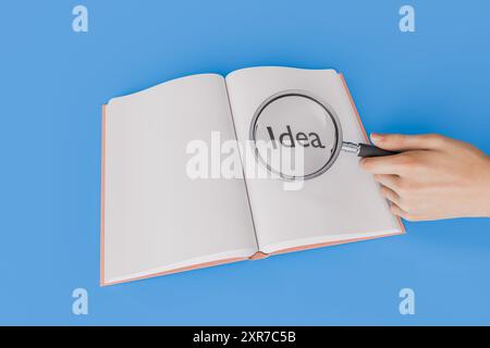 Eine Hand, die eine Lupe über ein offenes Buch hält und das Wort „Idee“ auf blauem Hintergrund vergrößert. Konzept von Innovation, Kreativität und RESEAR Stockfoto