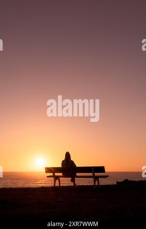 Frau, die auf einer Bank sitzt und den Sonnenuntergang am Strand genießt Stockfoto