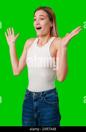 Junge Frau mit blondem Haar, die mit offenem Mund überrascht ist und die Hände gegen den Green Screen hochgehoben hat Stockfoto