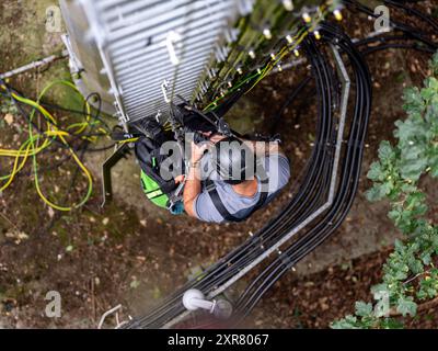 Telekommunikationsingenieur arbeitet in der Höhe, trägt einen Sicherheitsgurt und Helm und führt Wartungsarbeiten an einem Funkturm durch Stockfoto