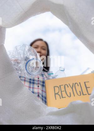 Asiatische Frau hält eine Kartonbox mit Recycling-Logo, die den Müll für separaten Müll zum Recycling sortiert und verschiedene Arten von Flaschen in den Müll wirft Stockfoto