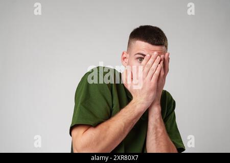 Mann, der Gesicht mit Händen in ängstlicher Pose vor grauem Hintergrund bedeckt. Stockfoto