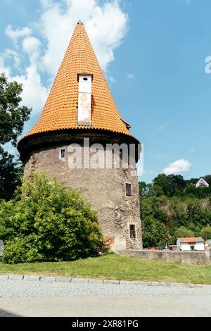 Ein runder Steinturm mit einem markanten orangen Dach steht majestätisch vor einem hellblauen Himmel. Um den Turm herum ist ein lebhaftes grünes Laub, enha Stockfoto