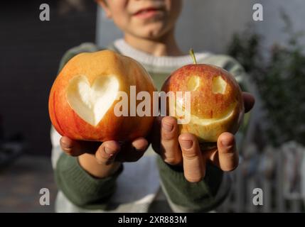 Das Kind hält zwei rote Äpfel in den Händen, auf dem einen ein Herz geschnitzt und auf der anderen ein lächelndes Gesicht. Richtige Vitaminernährung. Überraschung für Mama Stockfoto