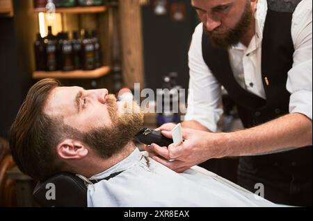 Professioneller Friseur mit Elektrorasierer zum Rasieren des Barts. Stammkunde bei einem Termin im Friseur. Selbstbewusster männlicher Kunde, der seinen Bart erledigt. Stockfoto