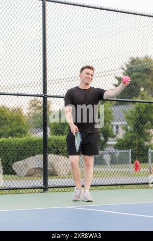 Junger Mann lächelt und hält einen rosafarbenen Pickleball und Paddel. Stockfoto