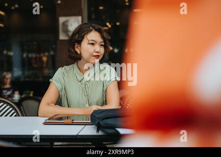 Nachdenkliche asiatische Frau in einem grün gestreiften Hemd, die mit einem Tablet an einem Kaffeetisch sitzt Stockfoto