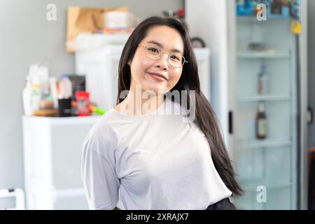 Eine schöne junge asiatische Frau, die in einem Haus steht. Sie lächelt hell und trägt eine Brille. Die Frau kann arbeiten oder sich zu Hause entspannen. Stockfoto