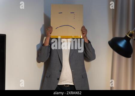 Unglücklicher Mann, der mit Box anstelle seines Kopfes arbeitet. Porträt eines Geschäftsmannes, der sein Gesicht mit Papiermaske bedeckt, mit traurigem Gesicht darauf gezeichnet, schlechte negative s Stockfoto