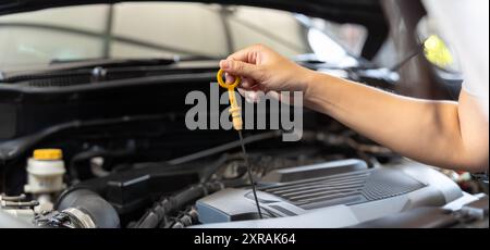 Frau, die das Motoröl im Schatten überprüft, arbeitet, das Motoröl überprüft. Frau, die sich um das Auto kümmert Check Auto Motoröl Konzept. Breites Bannerbild. Stockfoto