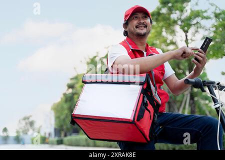 Ein glücklicher Liefermann sitzt auf einem Fahrrad und schreibt auf seinem Smartphone. Er trägt einen roten Rucksack zur Lieferung auf Schultern und Mütze. Das ist er Stockfoto