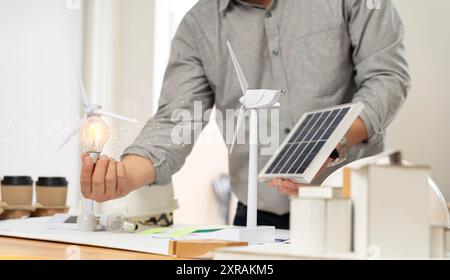 Nahaufnahme bei einer Glühbirne sitzt ein Ingenieur mit einer Glühbirne und einem Modell einer Windturbine. Mit Solarpaneelen und Modellhaus auf dem Tisch. Um u zu entwerfen Stockfoto