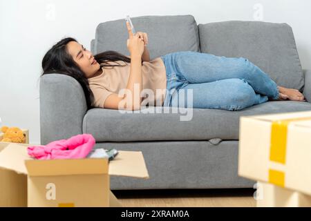 Frau, die das Smartphone benutzt, um auf das College zu ziehen, die Wohnung verlassen, um die Umzugswagen mit einem Paket Pappkartons für den Umzug in ein neues Haus in abzuholen Stockfoto