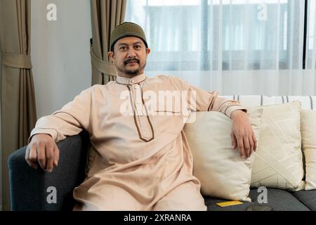 Porträt eines gutaussehenden jungen arabischen Mannes, der lächelt und in die Kamera blickt, zuhause auf dem Sofa sitzt. Attraktiver Mann aus dem Nahen Osten, der positive Emotionen ausdrückt. Stockfoto