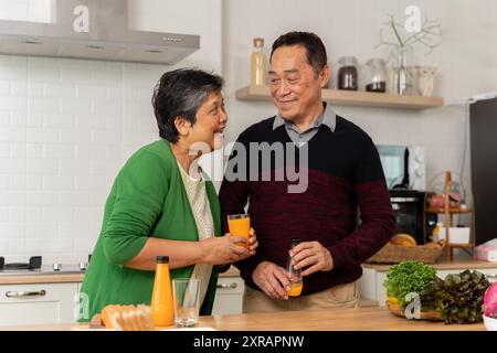 Liebevolles Seniorenpaar in Schürzen, sich gegenseitig mit Orangensaft toasten und gesundes Abendessen zubereiten, während Sie Zeit zu Hause verbringen. Verschiedene Früchte auf der Registerkarte Stockfoto