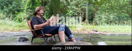 Lesen oder studieren Konzept Ein glücklicher bärtiger asiatischer Mann sitzt allein in einem Stuhl in der Nähe eines Baches auf dem Land und genießt die Waldlandschaft beim Lesen Stockfoto