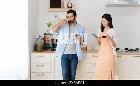 Junges Paar trinkt heißen Kaffee in der Küche mit Smartphone. Lateinische Frau lächelt und sieht den Freund an, während sie auf der Küchenzeile sitzt Stockfoto