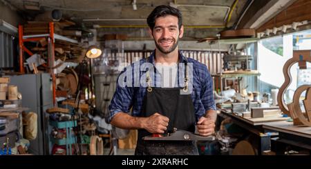 Herstellung von Lauten und Akustikinstrumenten. Mann mit kariertem Hemd, der Gitarre macht. Planen Sie die Gitarre zurück. Geigenwerkstatt. Dunkler schwarzer Hintergrund Stockfoto