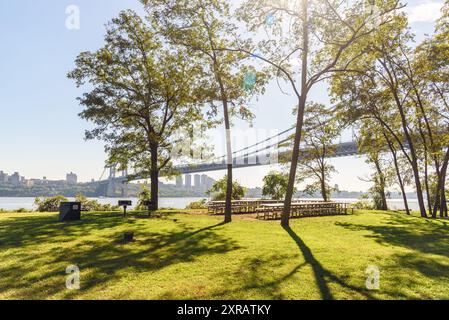 Picninc-Tische und Grillgerichte auf Gras zwischen Bäumen in einem Park am Ufer an einem sonnigen Herbsttag. Im Hintergrund befindet sich eine Lagen Supension Straßenbrücke. Stockfoto