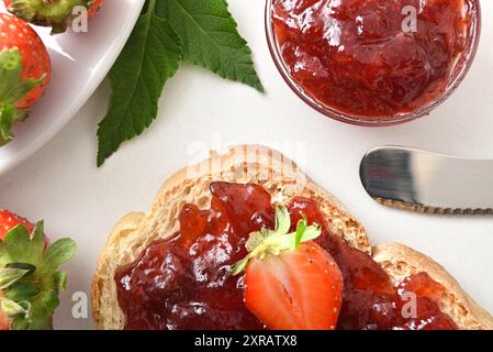 Detail der Erdbeermarmelade, die auf einer Brotscheibe und in einem Glasbehälter auf einem weißen Tisch mit Erdbeere und Messer verteilt wird. Draufsicht. Stockfoto