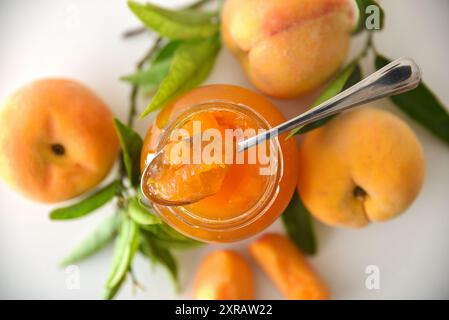 Glas mit Pfirsichmarmelade isoliert auf weißem Tisch mit Früchten und Löffel. Draufsicht. Stockfoto