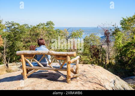 Eine Frau, die an einem sonnigen Herbsttag allein auf einer Holzbank auf einem Berg mit Blick auf das Flusstal eine majestätische Aussicht genießt Stockfoto