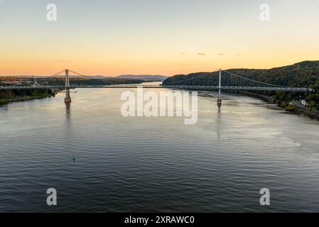 Sehen Sie sich die Hängebrücke an, die einen großen Fluss mit bewaldeten Ufern in der Abenddämmerung überspannt Stockfoto